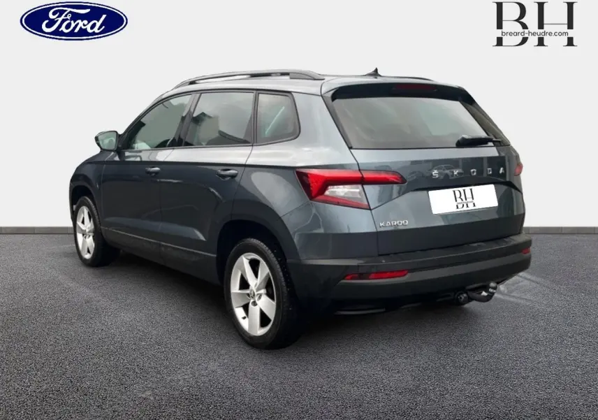 Vue 3/4 arrière droite d'un Skoda Karoq gris Business avec jantes alu et attelage visible sur fond neutre.