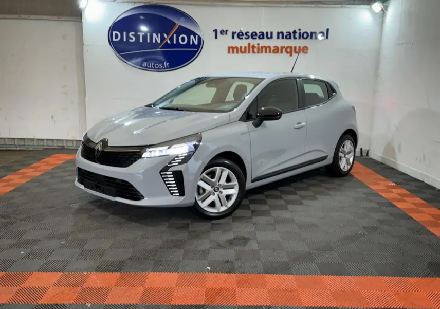 Renault Clio gris clair vue 3/4 avant droit, feux LED allumés, dans un showroom avec sol noir et orange.