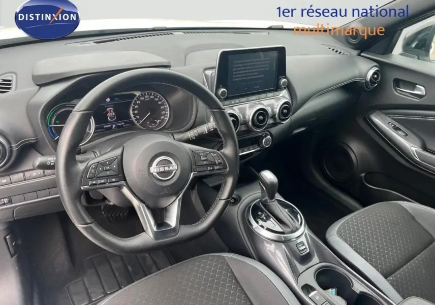 Vue intérieure avant du Nissan Juke blanc 2024, volant cuir, écran tactile et levier de boîte automatique visibles.