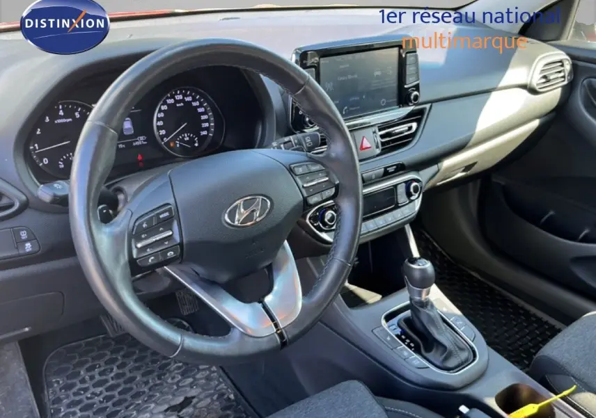 Vue intérieure du poste de conduite de la Hyundai i30 SW 2023, volant cuir noir et console centrale avec boîte automatique.