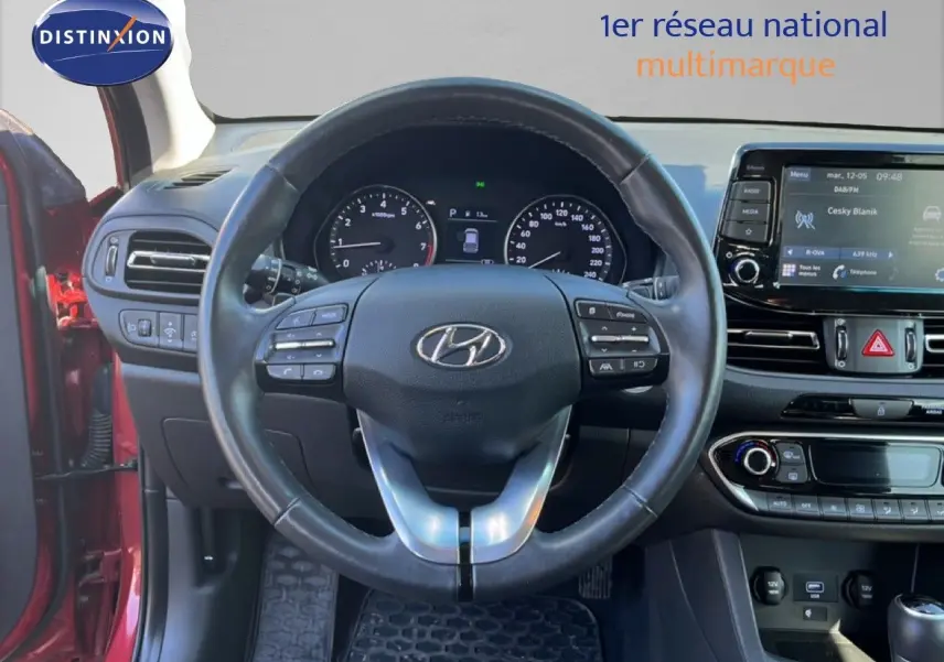 Vue intérieure centrée sur le volant et le tableau de bord d'une Hyundai i30 SW rouge avec écran tactile et commandes au volant.