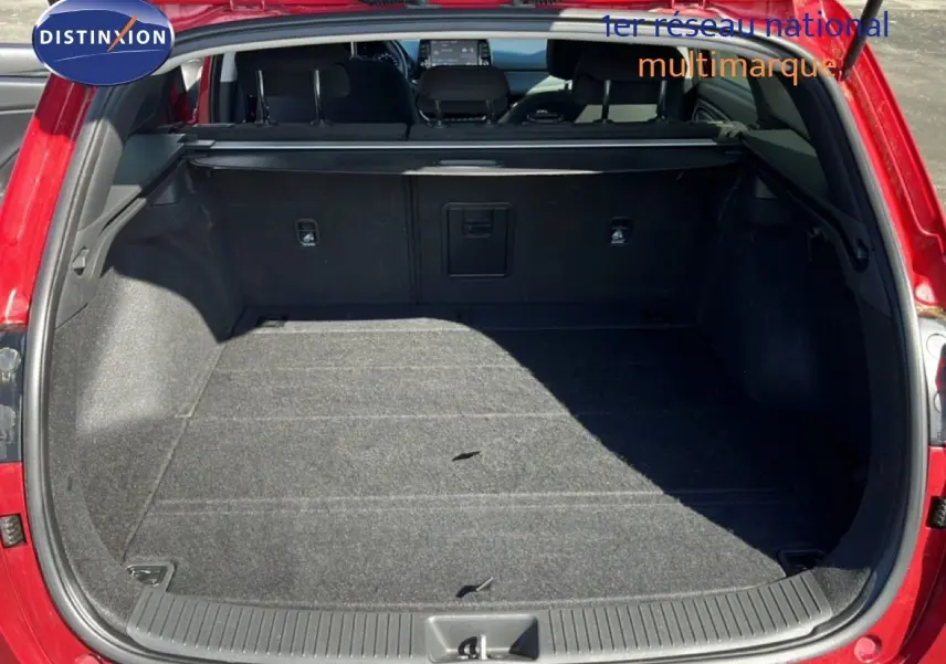 Coffre ouvert vu de l'arrière d'une Hyundai i30 SW 2023 en rouge Sunset Red Metal, intérieur noir et tapis gris.