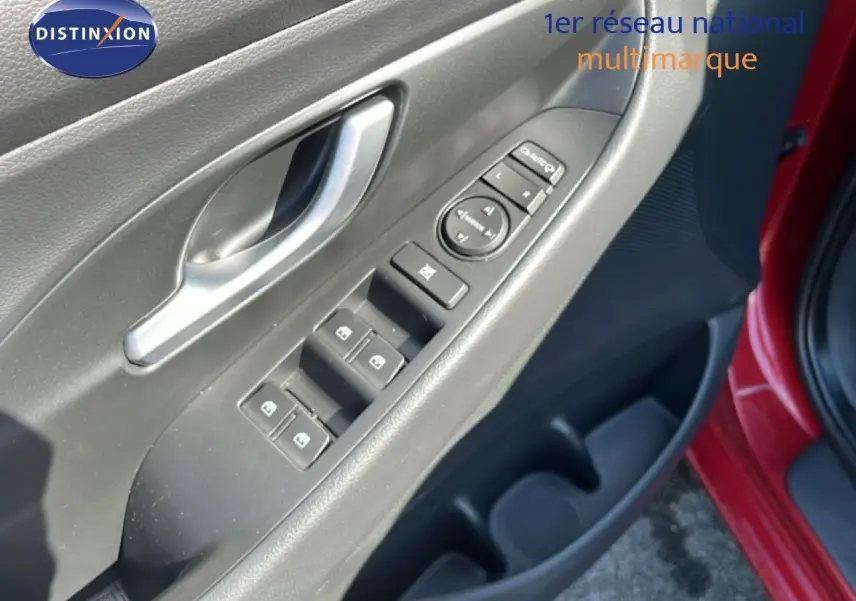 Gros plan sur la porte avant gauche d'une Hyundai i30 SW rouge, montrant les commandes des vitres et la poignée intérieure argentée.