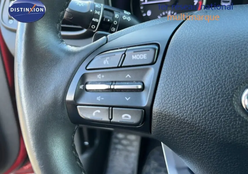 Gros plan sur le volant noir de la Hyundai i30 SW 2023, avec commandes audio et intérieur en rouge Sunset Metal visible en arrière-plan.