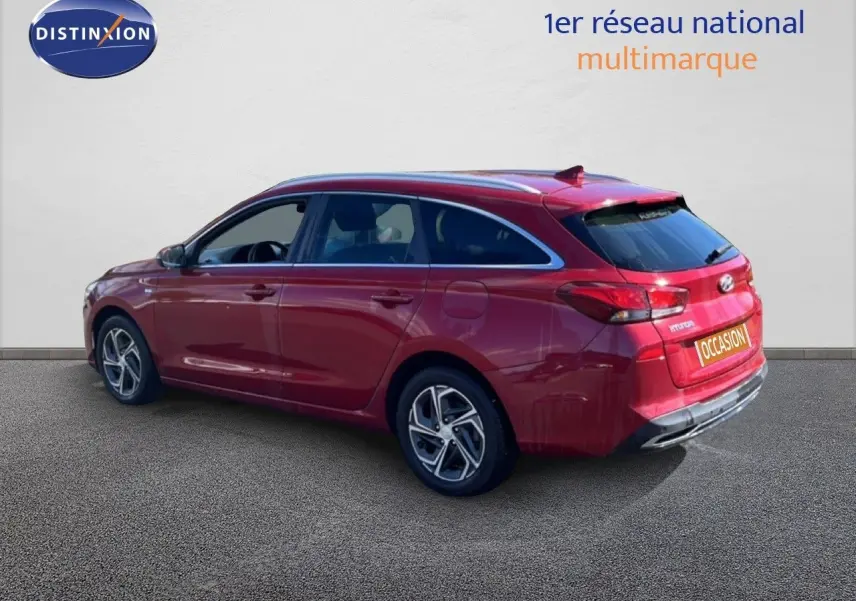Vue 3/4 arrière droite d'une Hyundai i30 SW 2023 rouge Sunset Red Metal avec jantes alliage et toit panoramique.