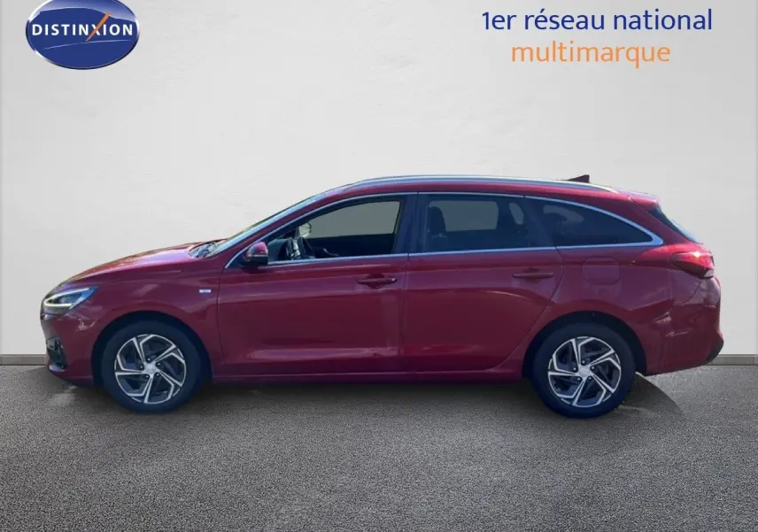 Vue de profil côté gauche d'une Hyundai i30 SW 2023 rouge Sunset Red Metal avec jantes bi-ton et vitres teintées.