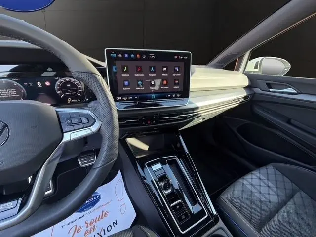 Vue intérieure côté conducteur de la Volkswagen Golf blanche 2025, mettant en valeur le tableau de bord digital et l'écran tactile central.