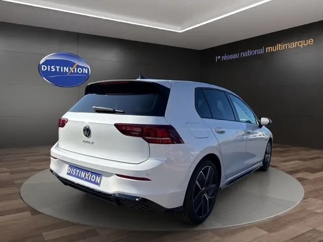 Vue 3/4 arrière droite d'une Volkswagen Golf blanche 2025 avec toit panoramique et jantes noires en exposition showroom.