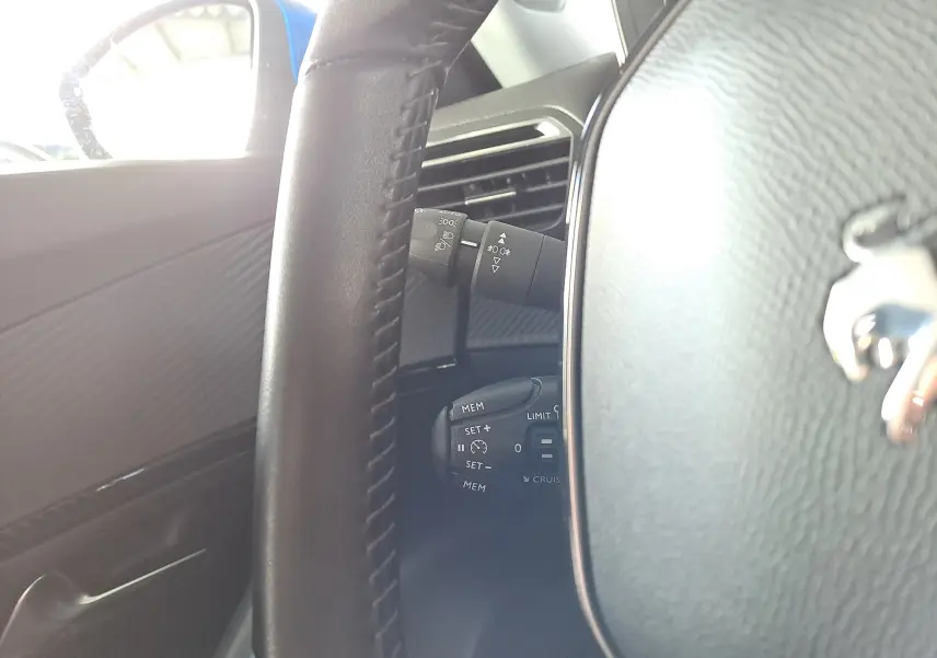 Gros plan sur le volant cuir noir du Peugeot 2008 bleu Vertigo, montrant les commandes de régulateur et lève-vitres côté gauche.