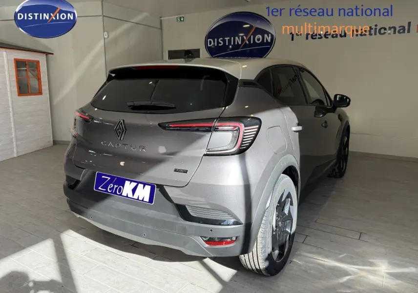 Vue 3/4 arrière droite du Renault Captur gris Cassiopée métal avec toit gris, en intérieur showroom, badge hybride visible.