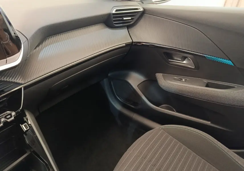 Vue intérieure côté passager de la Peugeot 208 2023, tableau de bord noir avec insert effet carbone et siège tissu gris.