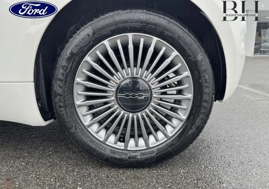 Gros plan sur la jante alu argentée d'une FIAT 500 Ice White pastel, roue avant droite visible sur sol humide.