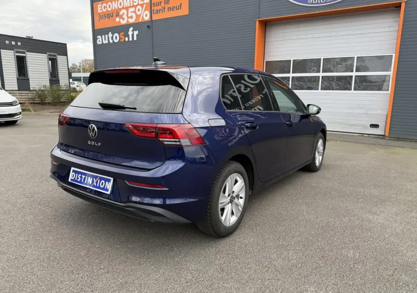 Vue 3/4 arrière droite d'une Volkswagen Golf bleu Atlantic métallisée avec vitres arrière surteintées, stationnée devant un garage autos.fr