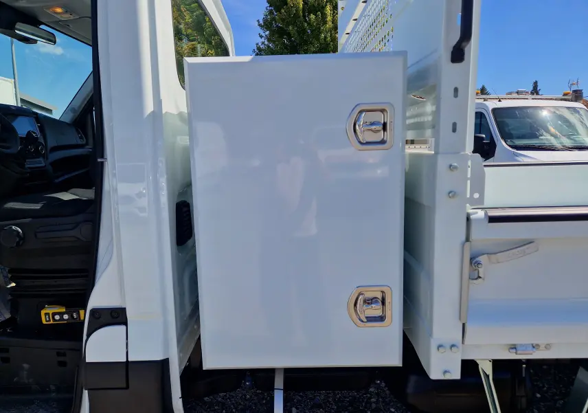 Vue latérale gauche rapprochée d’un Iveco Daily blanc montrant la benne acier avec coffre de rangement fermé.
