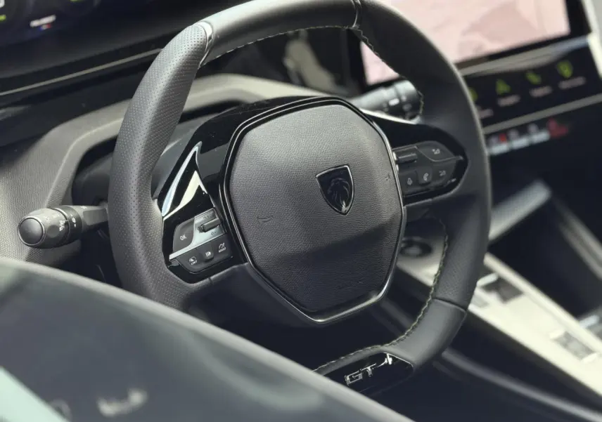Gros plan sur le volant cuir noir GT avec logo Peugeot et commandes intégrées, intérieur moderne de la 308 Hybrid 2026.
