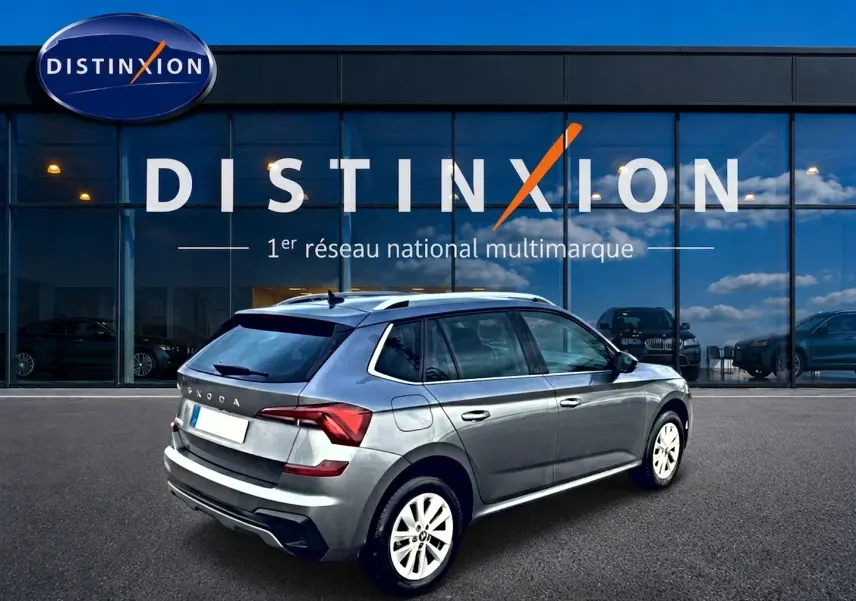 SKODA KAMIQ gris en 3/4 arrière droit, avec barres de toit et jantes alu blanches visibles devant un showroom Distinxion.