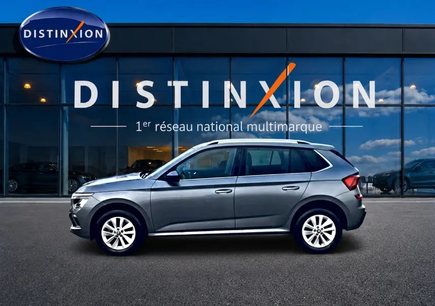 Vue latérale droite d'un Skoda Kamiq gris 2025 avec barres de toit et jantes alu devant une vitrine Distinxion.
