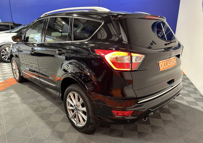 Ford Kuga noir en 3/4 arrière droit, avec feux allumés et jantes alliage dans un showroom.