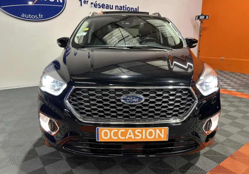 Vue frontale d'un Ford Kuga noir avec phares allumés et plaque "OCCASION" en orange, dans un showroom.