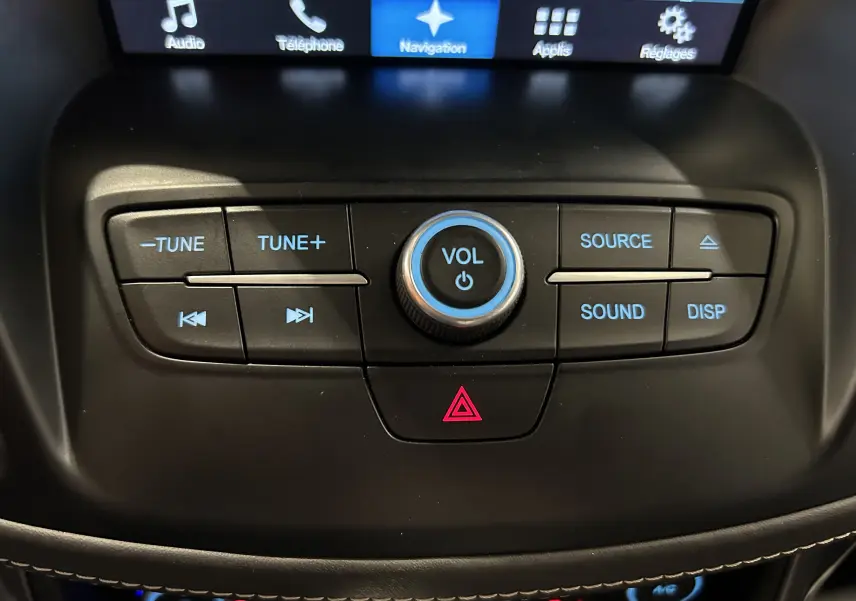 Gros plan sur la console centrale noire du Ford Kuga 2017, avec boutons de volume et commandes audio éclairés en bleu.