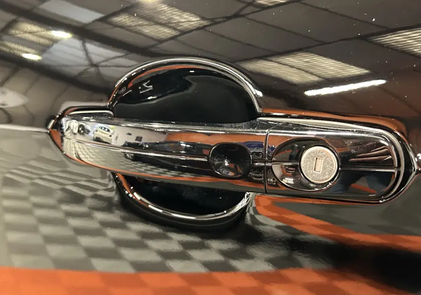 Gros plan sur la poignée de porte chromée avec serrure d’un Ford Kuga noir, reflet du sol damier orange et gris visible.