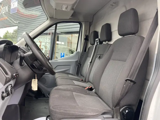 Vue intérieure côté conducteur du Ford Transit Fourgon blanc 2019, sièges tissu gris et tableau de bord visible.