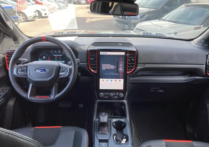 Intérieur avant du Ford Ranger Raptor 2026, vue du poste de conduite avec volant cuir noir et touches rouges, écran tactile central.