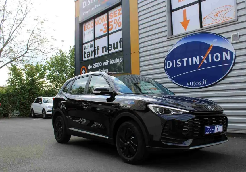 Vue 3/4 avant droit du MG ZS 1.5L HYBRID+ 2026 en noir Pebble Black Metal, stationné devant un bâtiment commercial.