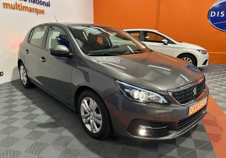 Peugeot 308 gris en 3/4 avant droit dans un showroom, avec phares allumés et plaque "OCCASION" visible.