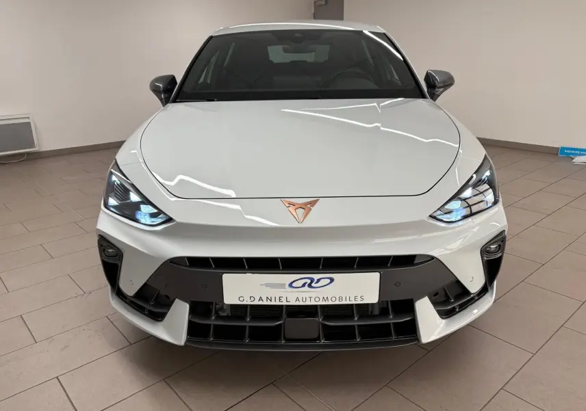 Vue frontale d'une CUPRA Leon Phase 2 blanche Nevada avec logo cuivré et phares allumés en intérieur.