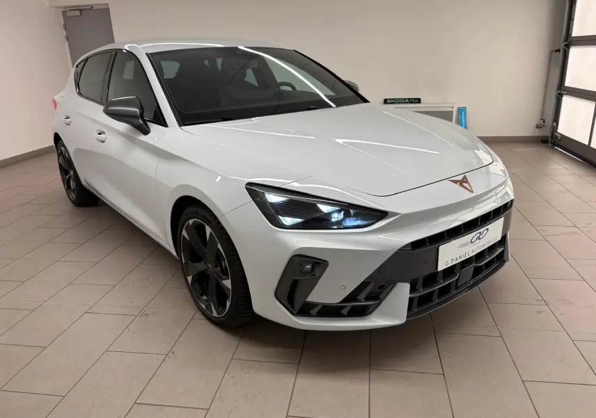 Vue 3/4 avant droite d'une CUPRA Leon blanc Nevada métallisé avec phares allumés et jantes noires distinctives.