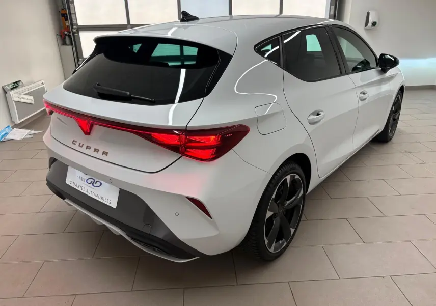 Vue 3/4 arrière droite d'une CUPRA Leon blanche Nevada avec feux arrière LED et jantes noires en intérieur showroom.
