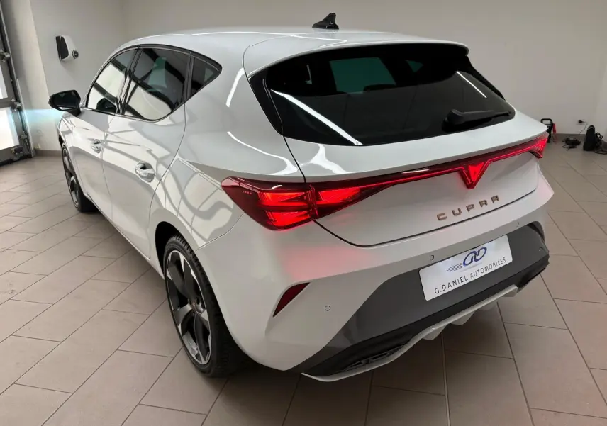 Vue 3/4 arrière droite d'une CUPRA Leon blanche Nevada avec feux arrière connectés et jantes noires en intérieur showroom.
