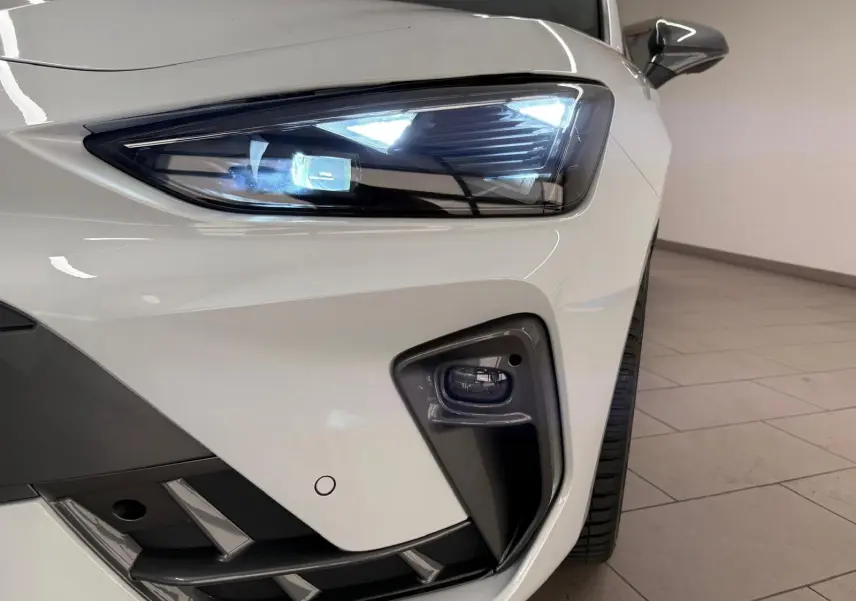 Gros plan sur le phare avant droit allumé d'une CUPRA Leon Phase 2 blanche Nevada avec antibrouillard intégré.