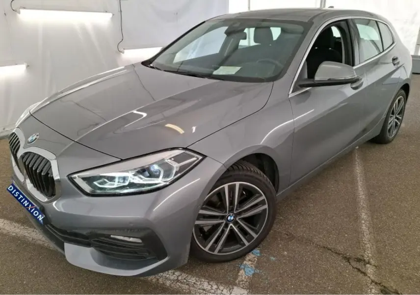 BMW Série 1 gris vue 3/4 avant droit, avec jantes alliage et phares LED, en intérieur showroom.