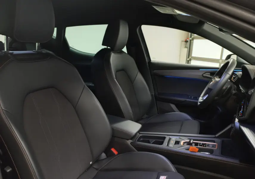 Vue intérieure côté conducteur de la CUPRA Leon 2025 avec sièges en cuir noir et éclairage d'ambiance bleu.
