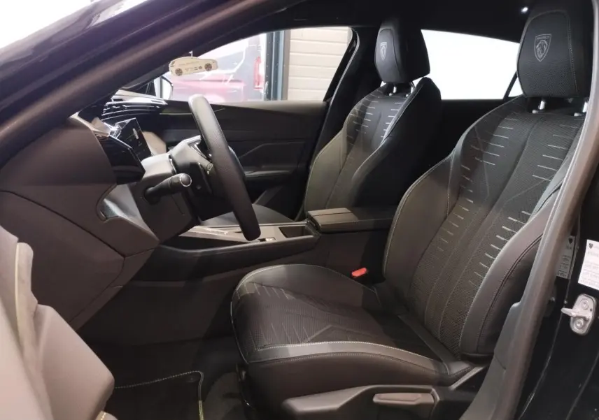 Vue intérieure côté conducteur de la Peugeot 308 Hybrid 2026 noire, sièges avec logo Lion et surpiqûres vert Adamite.