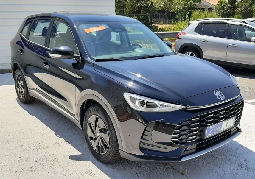 Vue 3/4 avant d’un MG ZS 1.5L Hybrid+ 197ch noir Pebble Black Metal avec calandre imposante et jantes aérodynamiques.
