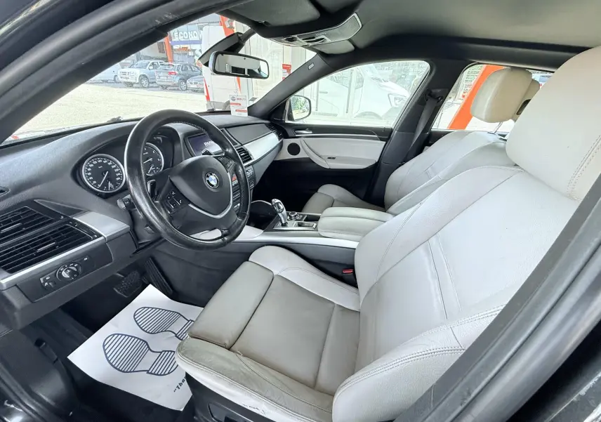Vue intérieure côté conducteur d’un BMW X6 2011 noir, sièges cuir clairs et tableau de bord noir avec volant multifonction.
