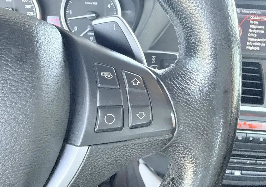 Gros plan sur le volant en cuir noir avec commandes intégrées, tableau de bord et écran central d’un BMW X6 noir de 2011.