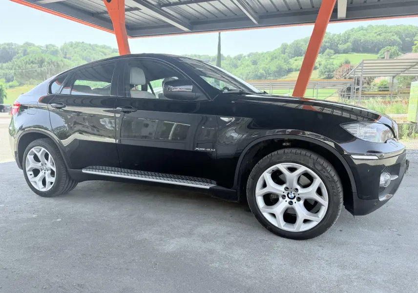 BMW X6 noire vue de profil côté gauche, sous un abri avec jantes alliage et marchepieds métalliques.