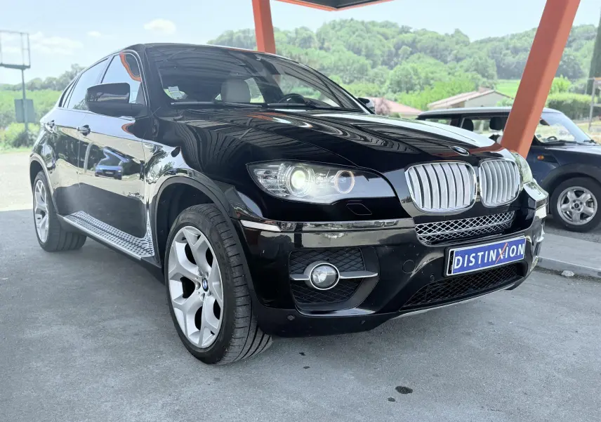 BMW X6 noire vue en 3/4 avant droit, avec calandre chromée et phares allumés sous un abri orange.