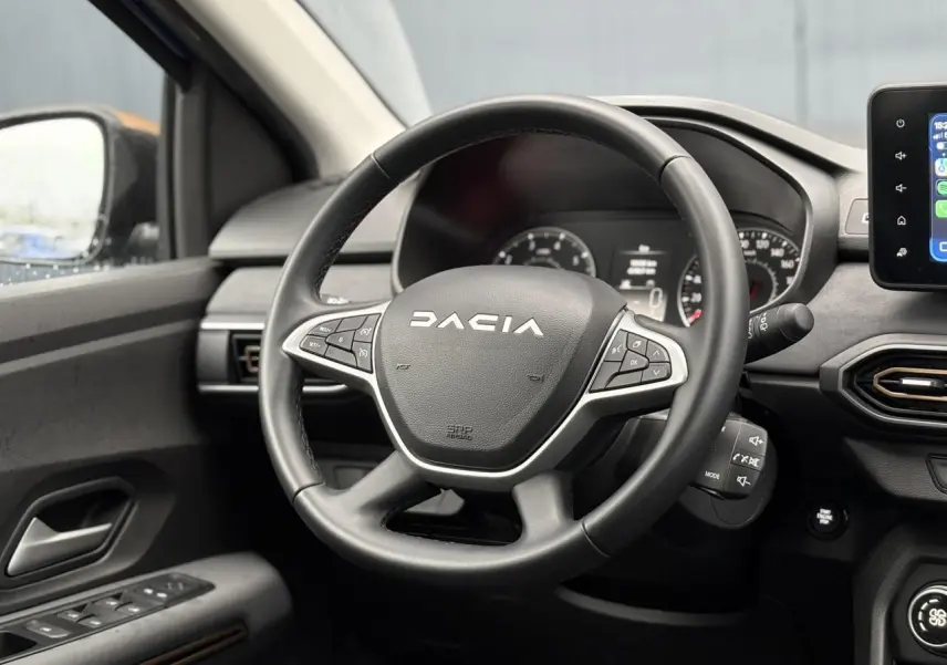 Vue intérieure centrée sur le volant noir Dacia avec commandes intégrées et écran tactile à droite.