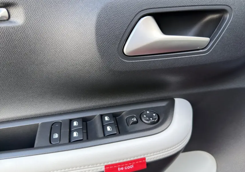 Détail de la porte côté conducteur avec commandes électriques et étiquette rouge "be cool" sur un intérieur noir et blanc.