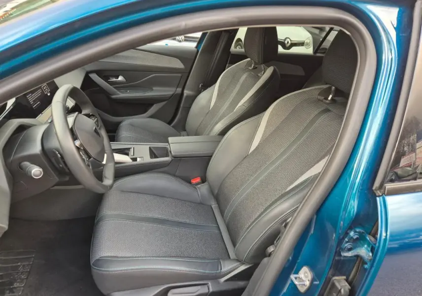 Vue intérieure côté conducteur d'une Peugeot 308 bleu 2024, sièges tissu et cuir avec accoudoir central et cockpit numérique.