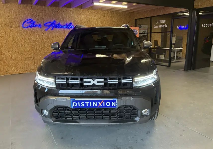Vue avant d'un Dacia Duster 1.6 Hybrid 140 noir, avec calandre distinctive et phares allumés en intérieur showroom.