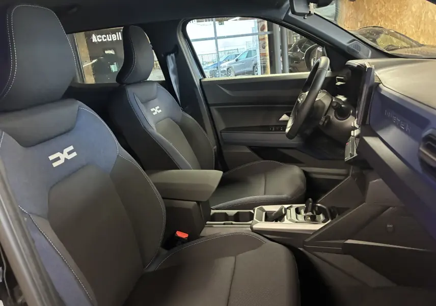 Vue intérieure côté gauche du Dacia Duster 1.6 Hybrid 140 Journey 2025, sièges tissu noir et gris avec logo Dacia brodé.