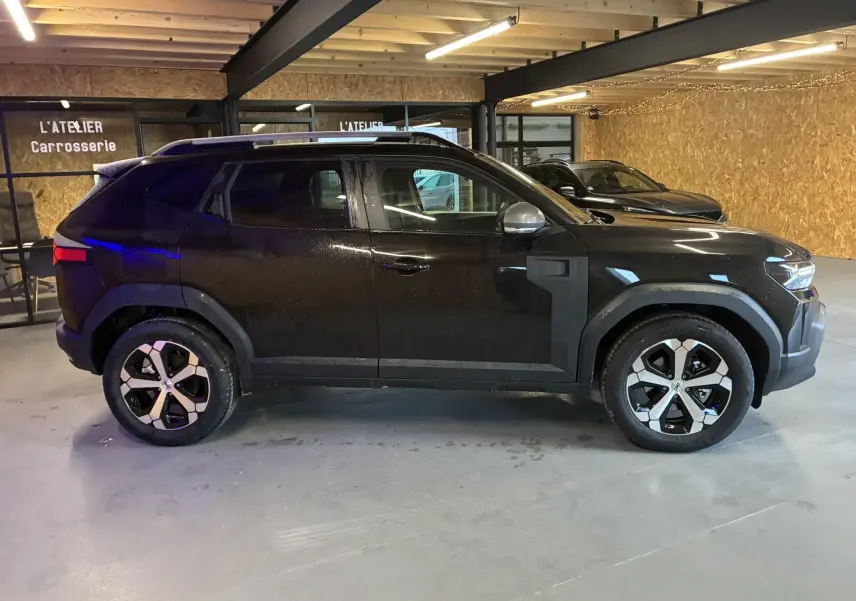 Profil droit du Dacia Duster 1.6 Hybrid 140 Journey noir, avec jantes distinctives et barres de toit, en intérieur d'atelier.