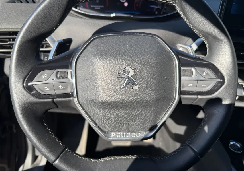 Gros plan sur le volant noir du Peugeot 5008 1.5 HDi 130 Allure 2021 avec commandes intégrées et logo lion central.