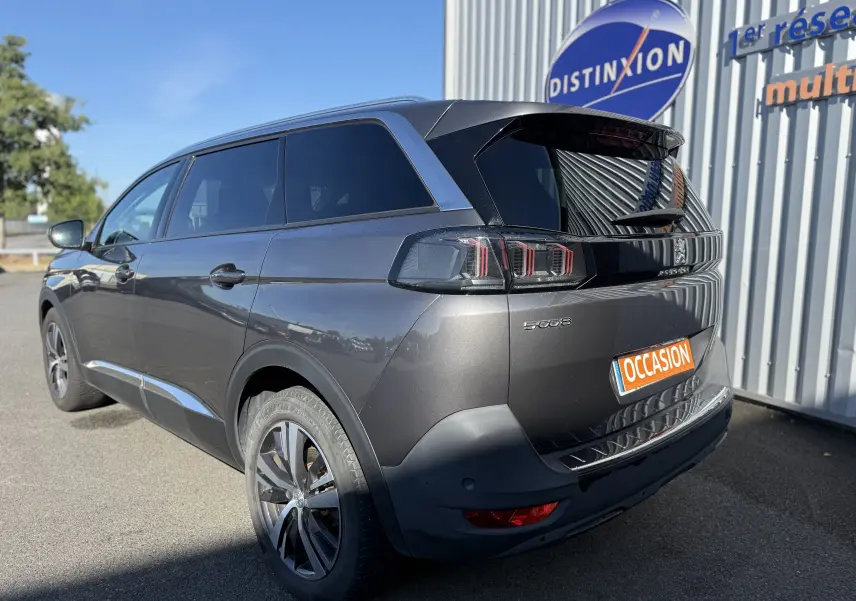 Vue 3/4 arrière droite d’un Peugeot 5008 gris métallisé avec jantes 18 pouces et vitres teintées, stationné devant un bâtiment.